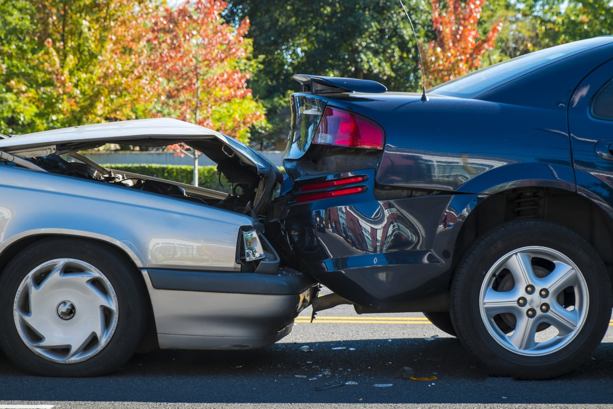 Two-car rear-end collision with visible damage to the front of a silver vehicle and the back of a dark blue vehicle on a road with trees in the background.