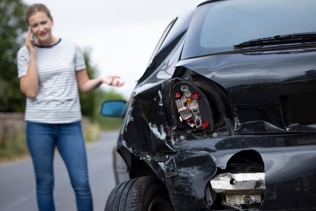 A woman on the phone stands beside a car with visible rear-end damage after an accident.