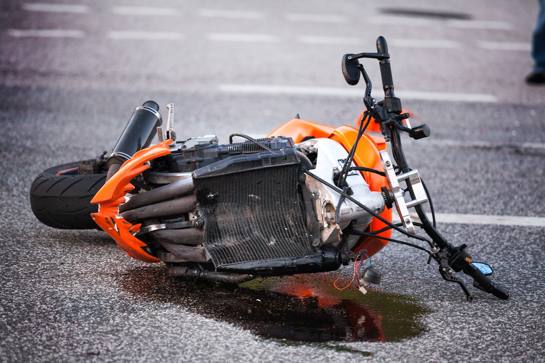 An orange motorcycle lying on the road after an accident, showing damage to the front and leaking fluid onto the pavement.