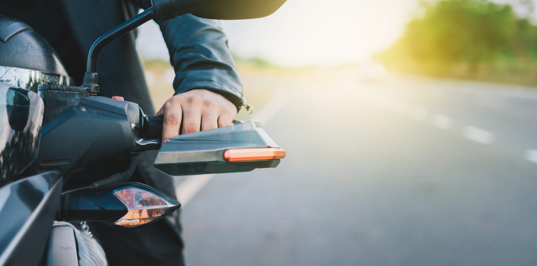 Close-up of a motorcyclist's hand on the handlebars, parked along a sunny open road.