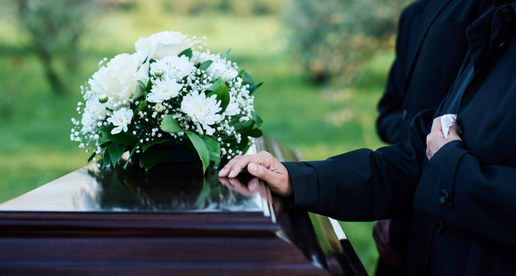 A mourner rests their hand on a casket adorned with white flowers during a funeral service.