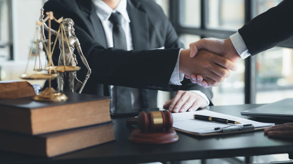 A lawyer and their client shaking hands across a desk with legal documents, scales of justice, and a gavel.