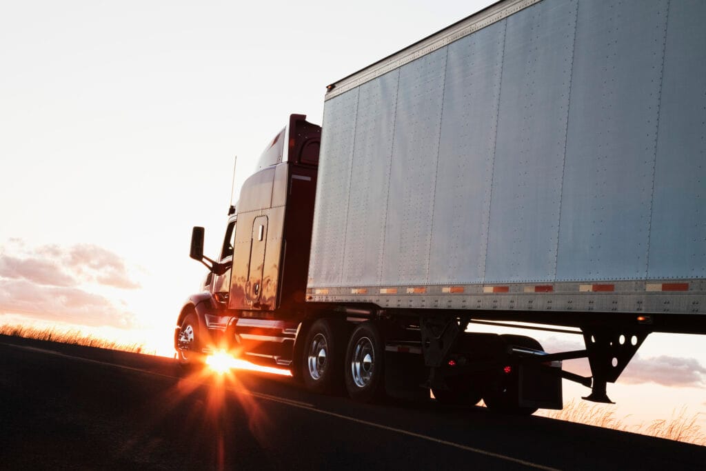 Silhouette of a commercial truck driving on a highway at sunset, with sunlight shining through the truck's wheels.